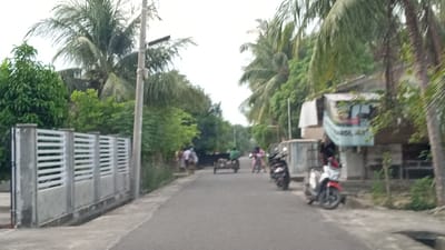 The image depicts a residential street scene in Cotgirek, Indonesia, characterized by lush tropical vegetation and modest dwellings. The road surface is asphalt, and on the left side of the frame, a modern concrete fence with horizontal slats runs along the property line. Coconut trees and other dense green foliage dominate the background, suggesting a warm climate.
Several people and vehicles are present on the road. In the mid-ground, a small cart is being pulled along the street, with an individual pushing it from behind. To the right of the cart, a person is riding a motorcycle. Another motorcycle is parked on the right side of the road in the foreground, and a person is seated on it. Further down the street, a few individuals are standing on the left side. A building with a banner is visible on the right, partially obscured by trees. The overall atmosphere is that of a typical day in a tropical village.