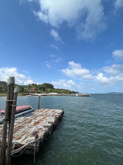 The image depicts a view from a pier in Pak Nam, Thailand. In the foreground, a weathered, rust-covered barge with several mooring ropes is visible, along with a portion of a boat with a red roof tied to it. The water is a deep green, with moderate waves. Beyond the barge, a long pier extends into the sea, lined with pilings and supporting various structures, including what appears to be buildings and a crane. A lush, green, tree-covered island rises in the background, with buildings nestled among the foliage. The sky is a bright blue, dotted with scattered white clouds, indicating a clear, sunny day. The overall scene suggests a coastal or harbor area, possibly a ferry terminal or a resort.