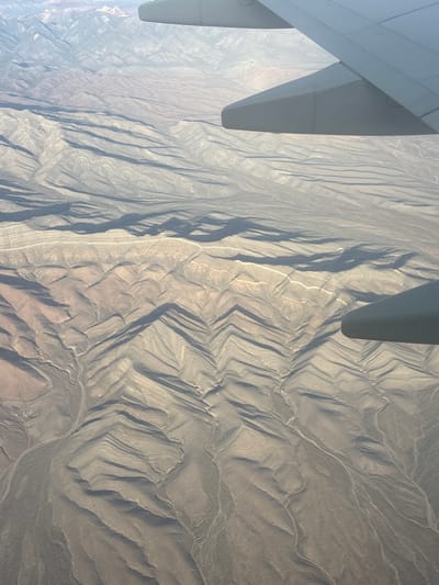 An aerial photograph depicts an expansive arid and mountainous landscape, observed from an aircraft. Portions of a light grey aircraft wing are visible in the upper left and right sections of the frame. The terrestrial environment below features numerous parallel geological ridges and valleys, creating a distinctive corrugated pattern across the terrain. Long shadows are cast by these formations, suggesting a low sun angle. The ground is characterized by shades of tan, light brown, and grey rock and soil, with minimal visible vegetation. Faint, winding light-colored lines indicate dry riverbeds or washes traversing the valleys. Hazy mountain ranges are present in the far distance. This landscape is consistent with the arid, mountainous desert regions surrounding Pahrump, Nevada, United States.