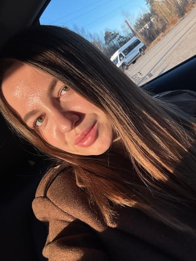A young woman is taking a selfie in a car. She is wearing a brown coat. Her hair is long and brown. She has light skin and brown eyes. In the background, a white van is driving on a road. The location may be Likino-Dulyovo, Russia.