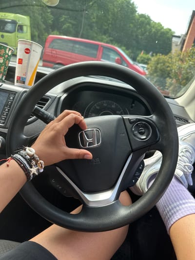 The image shows an individual in the driver's seat of a car, possibly a Honda, with the steering wheel prominently displayed. The person has a hand on the Honda logo. On the person's arm, multiple bracelets with shells and a starfish design are visible. The person is wearing lavender socks and white sneakers are visible near the top of the steering wheel. There is a glimpse of the person's bare leg. There are McDonald's cups on the dashboard. Outside the car window, there is a view of a green and a red van. This image does not provide enough information to determine if the photo was taken in Frechen, Germany.