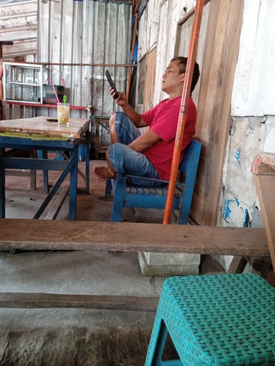 An adult man sits in a blue plastic chair, holding a smartphone. He is wearing a red t-shirt and blue jeans. The chair is located inside a simple structure, constructed from corrugated metal, boards, and plastic sheeting. The structure is possibly a small business. A blue table is on the left. A glass of beverage with two straws sits on the table. The foreground contains a bench, with a turquoise plastic stool underneath. This scene is located in Sekopek Wetan, Indonesia.