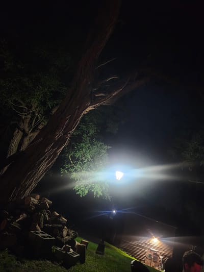 In the Town of Hoosick, United States of America, a nighttime scene unfolds under the illumination of a bright floodlight directed downwards and to the left. The light casts a strong glare, obscuring details in its immediate vicinity. To the left of the floodlight, a large tree trunk, textured and rough, dominates the frame, with its branches extending upwards and to the right. Leafy foliage is visible, illuminated by the light. In the foreground, a pile of cut logs is stacked on the ground, interspersed with concrete blocks. Further into the background, a wooden structure, possibly a shed or barn, is visible with its horizontally slatted walls and a doorway. The ground is covered in grass, which appears illuminated in patches by the light. The overall ambiance is that of an outdoor space at night, with artificial lighting playing a significant role in defining the visible elements.