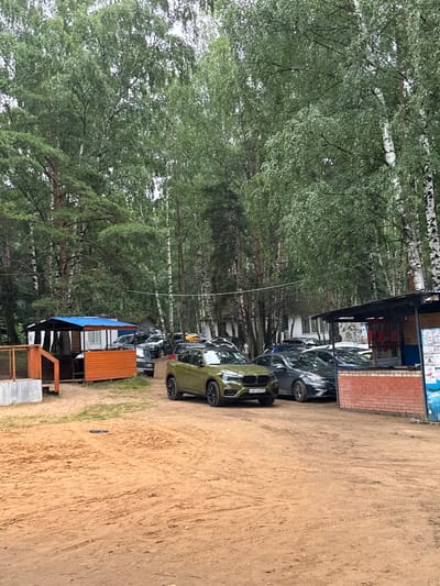 In a sandy, outdoor area surrounded by numerous tall trees, several vehicles are parked. In the foreground, a green SUV is visible, followed by a dark grey sedan. Behind these, a row of parked cars extends into the background, including a yellow car. To the left, a small structure with an orange wooden exterior and a blue roof is situated. Further left, a concrete structure with a wooden railing is present. A larger, covered area with a brick facade and a dark roof is visible on the right side of the image, appearing to be a kiosk or service area. The ground is covered in sand with visible tire tracks. The scene appears to be set in the Туристический Пансионат "Клязьминское водохранилище", Russia. There are no people clearly visible in the image.