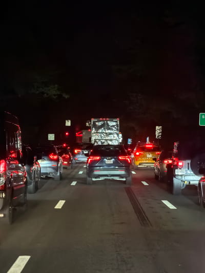 At night, a traffic jam is seen on a highway in Village of North Hills, United States of America. The vehicles are stopped in multiple lanes, with their brake lights illuminated. The surrounding environment is dark, with only the headlights and taillights of the cars providing illumination. A large truck with a reflective surface is visible in the distance, along with traffic signs. The road surface shows some tire marks.