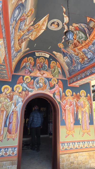 The image shows the interior of a church in Elenovo, Bulgaria. The walls and ceiling are covered in colorful Orthodox Christian iconography, depicting religious figures and scenes. A man stands in the doorway. He is wearing a blue jacket and dark pants. The doorway opens into the church's interior.