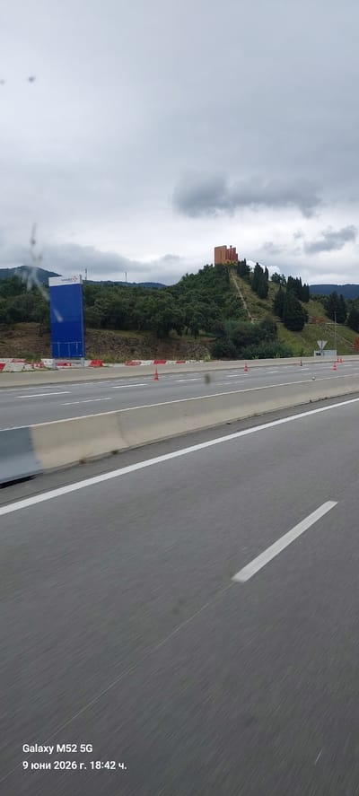 The image shows a view from a moving vehicle on a highway, likely near Le Perthus, France, given the provided timestamp in Cyrillic which includes "Le Perthus" and the date "9 June 2026". The scene features a multi-lane highway with concrete barriers and road markings, indicating a fast-moving environment. In the background, a hill covered with trees is visible, topped by a distinct red-orange structure that resembles a fortress or a modern building with fortress-like elements. To the left, a large blue sign with white lettering, possibly a company logo or directional information, stands against the backdrop of greenery and the overcast sky. Several orange traffic cones are scattered along the roadside, suggesting potential construction or lane closures. The overall impression is that of a typical highway landscape in a hilly region, with the unique architectural element on the hill serving as a focal point. The timestamp at the bottom left corner indicates the image was captured by a "Galaxy M52 5G" at "18:42".