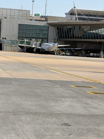 This image captures an outdoor scene at an airport, likely in Kempton Park, South Africa, given the provided location context. The primary subjects are an airplane docked at a terminal building and the surrounding tarmac.
The airplane, a commercial jet, is positioned next to a jet bridge, suggesting it is either boarding or deplaning passengers. Several airport service vehicles and a jet bridge are visible, indicating active operations. A few individuals, likely airport personnel, are present near the aircraft and vehicles, engaged in their duties.
The setting is an airport tarmac with clear markings on the ground, including yellow lines and rectangular shapes, which are typical for aircraft parking and movement areas. The terminal building is a modern structure with large glass windows, reflecting the sky and surrounding environment. The time of day appears to be daytime, with clear skies and bright sunlight casting distinct shadows. The overall atmosphere suggests a normal, operational day at a busy airport.
A notable detail is the text "A7" visible on a yellow sign near the jet bridge, indicating the gate number. The image is taken from a slightly elevated perspective, looking down onto the tarmac and the side of the airplane.