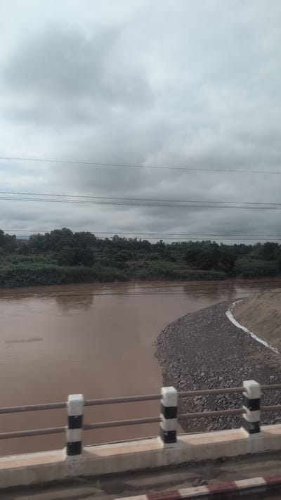 This is a medium-angle, landscape photograph taken from a bridge overlooking a wide, muddy river in Pakxan, Laos. The sky is overcast with heavy gray clouds. Several power lines run horizontally across the upper portion of the frame. The river, appearing brown and opaque, dominates the lower half of the image. On the right bank of the river, there is a constructed embankment consisting of dark grey gravel, with a strip of white material visible along its edge. The left bank of the river is densely covered with lush green vegetation, including trees and bushes, with a small, dark structure, possibly a hut, partially visible within the foliage. The foreground of the image shows the railing of the bridge, which is comprised of horizontal metal bars supported by concrete posts. These posts are decorated with alternating black and white stripes. The concrete structure of the bridge itself is visible below the railing, with a red and white striped barrier at the very bottom of the frame. The scene appears to be a natural landscape with some human infrastructure present.