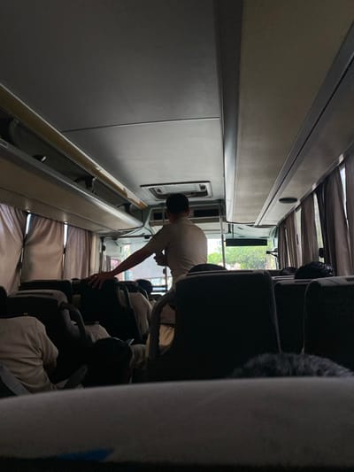 The image depicts the interior of a passenger bus in Boca del Río, Mexico. The perspective is from the rear of the bus, looking forward. The primary subject is a male individual standing in the central aisle, facing away from the camera. He wears a light-colored short-sleeved collared shirt and light-colored trousers. His right hand rests on the back of a dark-colored seat.
Multiple other individuals, primarily visible from the back of their heads and shoulders, are seated in the dark-colored, upholstered seats throughout the bus. Most visible passengers wear light-colored shirts or uniforms.
The bus interior features a light-colored ceiling with overhead compartments extending along both sides. Recessed lighting fixtures and an air vent are visible on the ceiling. Light-colored curtains cover the windows along the sides, partially obscuring the exterior view. Through the front windows, blurred green foliage and outdoor light are discernible, indicating the bus is in an outdoor environment. The lighting inside the bus is somewhat dim, contrasting with the brighter exterior light entering through the windows. No specific interactions are clearly defined beyond the standing individual's posture and proximity to seated passengers.