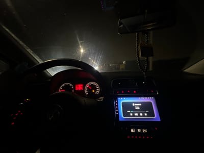 The interior of a car is shown at night. The steering wheel and dashboard are visible, with illuminated gauges. The central console features a screen displaying music playback controls. A rearview mirror hangs from the ceiling with a decorative object suspended from it. The scene is dimly lit, with light sources appearing to be the car's instruments and exterior streetlights. The overall impression is a nighttime driving environment.