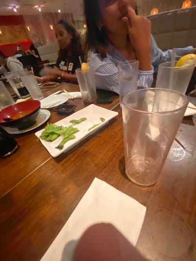 This image depicts the interior of a commercial dining establishment, consistent with a restaurant, potentially located in West Windsor, United States, though the exact venue name or address cannot be determined from the visual information provided.
In the immediate foreground, a human finger is partially visible at the bottom right, slightly obscuring a white folded napkin. An empty, clear plastic drinking glass rests prominently on a dark, polished wooden table surface, exhibiting visible water rings.
On the table in the mid-ground, a white rectangular dish contains several green edamame pods and some discarded empty shells. To its left, a black bowl with a red interior rim holds an orange-colored liquid, likely soup, positioned on a white ceramic plate. A smaller, empty white ceramic plate, small black square dishes, and multiple clear drinking glasses are also present. Some glasses contain clear liquid, at least one with a lemon slice and a straw. A pair of chopsticks rests on the table. Two mobile phones are visible on the table surface.
Two individuals are primarily visible. On the right, a person wearing a light blue and white striped shirt is seen from the chest up, with their right hand positioned near their face, touching the nose area. Further back on the left, another person with long dark hair, wearing a dark shirt with partially legible white text ("EN HOME"), is seated and looking towards the left. Other blurred figures are seated at tables in the background.
The background shows a dimly lit restaurant interior featuring red wall elements, light-colored booth seating, and hanging light fixtures, creating a modern casual dining ambiance.