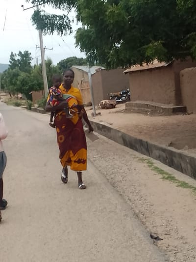 A woman is carrying a child along a dirt road in what appears to be a rural area of Biliri, Nigeria. The woman is wearing a yellow top and a long, patterned skirt of orange and red. She is wearing sandals and holding a piece of paper in her free hand. The child is wearing a colorful patterned outfit. The road is unpaved and there is a concrete drainage ditch running alongside it. In the background are simple, mud-walled buildings with corrugated metal roofs, some trees, and a power line. A motorcycle is parked outside one of the buildings. Another person's arm and shoulder appear in the lower left corner. The sky appears overcast.