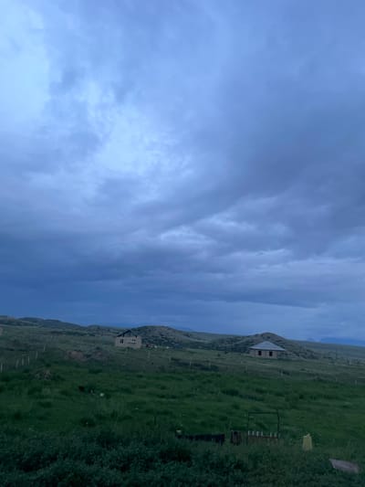 This is an outdoor, eye-level, full-shot photograph taken in the city of Kara-Oi, Kyrgyzstan, depicting a rural landscape under a cloudy sky. In the foreground, tall green grass and bushes are visible, with some domestic items scattered among them including what appears to be laundry hanging on a line, a wooden fence, and a yellow plastic bag. A dark object, possibly an animal, is in the mid-ground. Further back, two houses are situated on rolling green hills. The house on the left is lighter in color and appears to have windows and a dark roof. The house on the right has a pitched roof and visible windows. The landscape is characterized by undulating hills and a muted color palette, suggesting it may be dusk or dawn. The sky is filled with heavy, dark clouds, indicating potential for rain or stormy weather.
