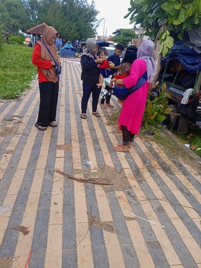 The image shows a street in Pameungpeuk, Indonesia (latitude: -7.668, longitude: 107.694). There are five people standing on a striped concrete walkway, composed of alternating tan and gray tiles. The tiles are damaged and cracked. Four of the people are women wearing headscarves. The person at the front is wearing a pink headscarf and a long pink dress. She is holding a young child in her arms, wrapped in a blue fabric. The woman behind her is wearing a gray headscarf, a long blue shirt, and long black pants. She is holding a young child in her arms. The woman behind her is wearing a blue headscarf and a long black shirt.  The woman at the back is wearing a red shirt, a blue headscarf, and black pants. She is carrying a dark purse and is holding a black bag. The fifth person is a man wearing a blue shirt and black pants. He is standing behind the first woman and next to the woman wearing the gray headscarf.  There is a large truck to the right side of the image that is obscured by vegetation and other vehicles.  The ground is covered in dirt and some debris.  The image is taken from a slightly high angle.  The sky is overcast.  The setting is a rural area near a beach.  The people appear to be casual, but are interacting in a focused way.  The children are being held and attended to by adults.  There is no indication of what the subjects are doing, but they are likely taking a break from their day and interacting with each other.  The scene is peaceful and calm.