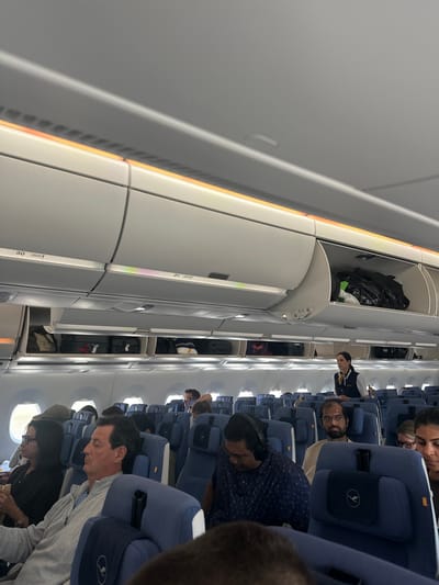 An indoor, eye-level, wide shot captures the interior of an airplane cabin, likely en route to or from Freising, Germany, given the context of Lufthansa's presence. The overhead compartments are open, revealing luggage, primarily suitcases, stored within. Passengers are seated in blue upholstered seats, some with headrests featuring the Lufthansa logo. A female flight attendant, wearing a navy uniform with a yellow scarf, stands in the aisle, appearing to be interacting with passengers or attending to duties. Several passengers are visible, some looking forward, others engaged in personal activities. The lighting suggests the cabin is illuminated by overhead lights, with warm-toned ambient lighting visible along the top edge of the cabin. The overall scene depicts a typical commercial flight environment.