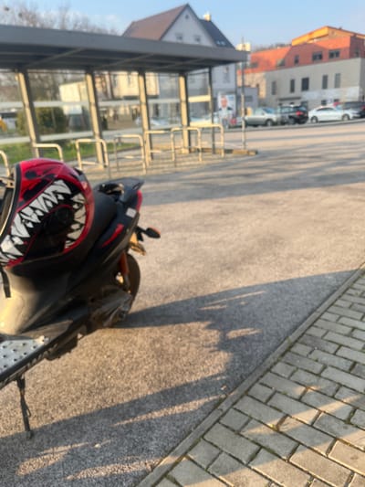 A black scooter with orange suspension accents is parked on its kickstand on a paved surface, facing towards the left. A red and black full-face helmet, featuring a prominent white, toothy monster mouth graphic, rests on the scooter's seat. Long shadows are cast across the asphalt from the right, indicating low-angle sunlight. To the right, a light-colored brick sidewalk runs diagonally. In the midground, behind the scooter, a modern bus stop shelter with a grey frame and glass panels is visible, with multiple metal bicycle racks positioned in front of it. In the background, various buildings with pitched roofs, some light-colored and one with a reddish-orange roof, line a street or parking area where several cars are parked. Bare trees are visible to the far left. A political poster with a man's face is affixed to a pole in the background. No individuals are present in the scene. The specific location within Krško, Slovenia, is not identifiable from the image alone.