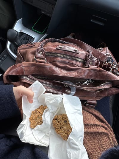 The scene depicts the interior of a vehicle in Kliczków, Poland. A light-skinned left hand, adorned with a dark blue sleeve, holds two individual oatmeal-style cookies, each wrapped in crinkled white tissue paper or napkins. The cookies contain visible ingredients such as oats, nuts, and possibly dried fruit. Positioned to the right of the hand is a distressed brown leather satchel-style handbag, partially open, featuring multiple exterior zippers, braided handle details, and metal hardware. Below the handbag, a textured brown knitted garment, possibly a scarf, is visible. In the upper left background, elements of the vehicle's dashboard are present, including a black automatic gear selector, silver trim surrounding the center console, and a storage compartment. Within this compartment, a black smartphone encased in a bright green protective cover rests, with USB-A and USB-C charging ports visible above it.