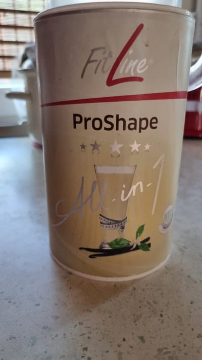 An up-close, eye-level shot shows a beige cylindrical container with the "FitLine ProShape All-in-1" branding prominently displayed. The logo "FitLine" is in a stylized font with a red accent line, and "ProShape" is in a bold, dark font. Below "ProShape" are three stars of varying sizes. A glass of a pale yellow beverage, possibly a shake, is depicted on the front, with a measuring tape wrapped around it. Vanilla beans and leaves are also illustrated on the container. A small circular logo with "NTC" is visible on the right side. The container is placed on a speckled, light-colored countertop, which appears to be in a kitchen or home environment. In the blurred background, a window with horizontal blinds and a glimpse of a white object are visible. The lighting suggests an indoor setting, possibly with natural light coming from the window.