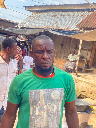 This image depicts an outdoor street or market scene in Igbara, Nigeria, under bright daylight. The central figure is a dark-skinned adult male facing directly forward with a serious expression, a slightly furrowed brow, and a small, slender object (possibly a toothpick or plant stem) positioned between his lips. He wears a long-sleeved green t-shirt with orange trim at the collar and cuffs, featuring a prominent graphic print of multiple champagne bottles, including visible "MOET" branding and a circular "PREMIUM" seal.

To the left of the central figure, a dark-skinned adult male is partially visible, wearing a light-colored button-up shirt and a beard, looking towards the left of the frame. In the background, other individuals are present; one person wearing a white t-shirt and light-colored pants is seen from the back. To the right, an older dark-skinned adult female is seated on a low blue stool against a wooden plank structure. She wears a light-colored button-up shirt, a patterned skirt, and a patterned head covering, with her hands clasped in her lap.

The surrounding environment consists of various structures characterized by corrugated metal roofs, some exhibiting significant rust and weathering, indicative of age and exposure. Walls appear to be constructed from wooden planks or other simple materials. Multiple tarpaulins or fabric canopies, secured by ropes, are stretched overhead, providing shade over parts of the area. The ground is uneven, composed of packed earth or sand, with some visible concrete or stone elements forming steps or platforms. Objects such as black plastic basins/bowls, sacks, and miscellaneous debris are scattered on the ground and on elevated surfaces. The upper portion of the image reveals a partially cloudy blue sky.