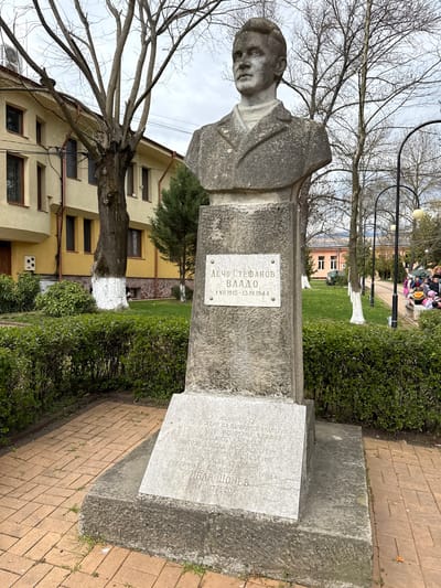 The image displays a stone bust of a man, mounted on a concrete pedestal, located in an outdoor park-like setting in Chavdar, Bulgaria. The bust depicts a young man with short hair, facing slightly to his left. The day is overcast, and the lighting is diffused, indicating daylight.
The pedestal features two white marble plaques. The upper plaque identifies the honored individual as "ДЕЧО СТЕФАНОВ ВЛАДО" (Decho Stefanov Vlado) with the lifespan "1 VII 1915 - 13.IV.1944". The lower, larger plaque commemorates a historical event: "НОЕМВРИ 194" (November 194X, likely 1943 or 1944, given Vlado's death year). It states that "В ЧЕСТ НА ДЕНЯ НА НАРОДНИТЕ БУДИТЕЛИ" (In honor of the Day of National Awakeners), "ЧЕТА БАЧО КИРО ОТ ОТРЯД ЧАВДАР" (Baсho Kiro's detachment from Chavdar) "ОБАДИЯ СЕЛО ЧАВДАР" (called upon Chavdar village). The text further describes that "НА ТОЗИ ПЛОЩАД БЯХА ИЗГОРЕНИ ДАНАЧНИТЕ КНИГИ И СЕ СЪСТОЯ ОБЩОСЕЛОКО СЪБРАНИЕ" (On this square, the tax books were burned, and a general village meeting took place). It concludes with "ГОВОРИ ЗАИ КОМАНДИРА НА ОТРЯДА - ИВАН ШОНЕВ" (spoke the commander of the detachment - Ivan Shonev), followed by "ДМИТРЕТО". This plaque details an anti-fascist action during World War II.
In the background, to the right of the bust, there is a paved brick path, leading to a grassy area with neatly trimmed bushes and bare-branched trees. Streetlights are visible along the path. Further in the background, a group of people, possibly children and adults, can be seen near a bench, suggesting a public park or square. To the left, a yellow multi-story building with multiple windows is visible, partially obscured by the bare branches of a large tree. The base of some tree trunks in the park are painted white.