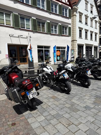 The image depicts a street scene in Riedlingen, Germany, featuring a row of parked motorcycles in front of a white building with distinctive architectural details. The building has multiple rows of windows; the upper windows have horizontal louvers, while the lower windows have green shutters. Banners with the words "Kultur Werk Stadt" and "stille cha" hang near the entrance of the building. The foreground is dominated by several motorcycles, including a red BMW model with registration plate "BC E 398." Other motorcycles, mostly black and white, are parked behind it, with license plates "KN LB37" and "LB92." The street is paved with rectangular stone blocks.  A sign near the building entrance mentions "ARAOKE".