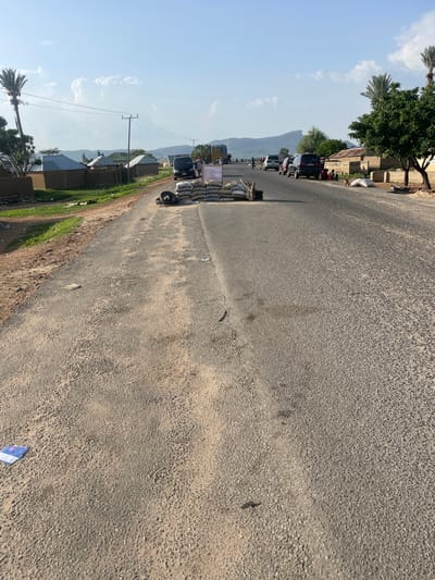 The image depicts a roadside scene in Wai A, Nigeria, with a road running through a semi-urban or rural area. In the foreground, the asphalt road is visible, showing signs of wear and tear. Several sandbags are piled up on the side of the road, potentially indicating a checkpoint or temporary barrier. A couple of old tires are also present near the sandbags.
On the left side of the road, a dirt path leads to a cluster of buildings that appear to be traditional houses with mud walls and corrugated metal roofs. Palm trees and other vegetation are scattered around the buildings. A utility pole with power lines stretches across the scene.
Further down the road, several vehicles are parked or moving. These include vans, SUVs, and possibly trucks, suggesting ongoing traffic. A few individuals are visible near the vehicles and on the side of the road, some appearing to be sitting or standing. One group of people is gathered near the buildings on the right. The background features a hilly or mountainous landscape under a bright sky with scattered clouds. The overall impression is of a typical day in a Nigerian community, with elements of daily life, infrastructure, and the natural environment.