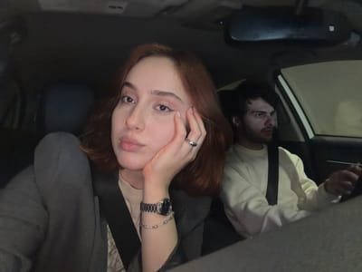 Two individuals are inside a vehicle. A person with reddish-brown hair is resting their hand on their face, wearing a watch and a bracelet on their left wrist. Their ring finger has a ring on it. A second person is visible in the background, wearing a light-colored shirt and a tie. The interior of the car is dimly lit. The location is not identifiable as Grigalati, Georgia.