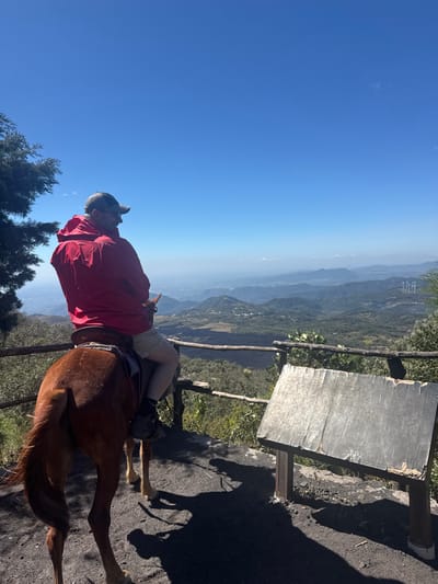 A man wearing a red jacket and hat is riding a brown horse. The horse is standing on a dirt path next to a wooden fence and a weathered wooden sign. Behind the fence, there is a view of a valley with green trees and mountains in the distance. The sky is blue and clear. The sun appears to be shining brightly. The photo is slightly rotated to the right.