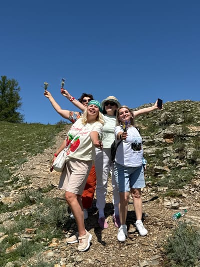 Four individuals are posing for a photograph in a mountainous, outdoor environment with a clear blue sky overhead. The scene appears to be a hiking trail in the Bichiktu-Boom Gorge, Russia, characterized by rocky terrain, sparse green vegetation, and a large tree on the left.

The individuals are standing on a dirt path. The person in the foreground, on the left, is a woman with blonde hair, wearing a white t-shirt with a strawberry graphic and the words "STRAWBERRY MIST", beige shorts, and light beige sneakers. She is holding a small bouquet of wildflowers in her right hand and a white purse on her left arm.

To her right is another woman with blonde hair, wearing a light green long-sleeved shirt, white pants, and pink sneakers. She is wearing a straw hat and sunglasses, and holding a phone in her outstretched right hand, possibly taking a selfie. She is also holding a small bouquet of wildflowers.

Behind and to the left of the first woman is a third person, whose gender is difficult to determine, wearing a colorful floral shirt and holding a small bouquet of wildflowers.

The fourth individual is partially visible behind the other three, wearing a white t-shirt with a graphic that includes text and an image of a mountain. This person is also holding a smartphone.

The overall atmosphere is casual and celebratory, with the individuals appearing to be enjoying an excursion. A plastic water bottle is visible on the ground to the right.