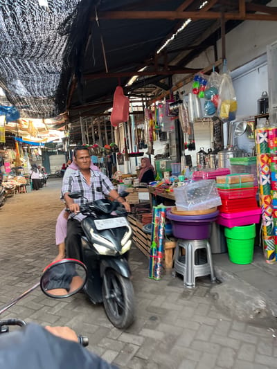 The image depicts a bustling outdoor market scene in Gedang, Indonesia. In the foreground, a person is riding a motorcycle, their hand visible on the handlebar, with a rearview mirror reflecting their legs. The motorcycle is black with a white license plate. The rider, a man with a checkered shirt and dark pants, is driving. Behind him, a woman in a light pink headscarf and dress is seated as a passenger.

The market environment is characterized by narrow pathways paved with brick-like stones. On either side of the path, stalls are filled with a variety of goods. These include an abundance of plastic containers in various colors and sizes, cookware, and what appear to be bundles of colorful items, possibly decorations or goods wrapped in plastic. Some stalls have awnings or tarps overhead, providing shade. In the background, other market-goers and motorcycles can be seen, indicating a lively commercial atmosphere. The overall lighting suggests it is daytime, likely morning or afternoon.