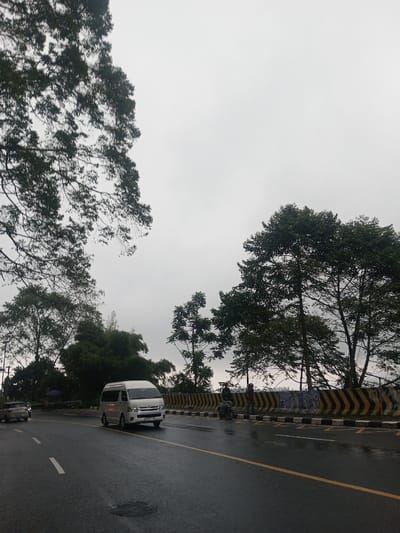 The image captures a road scene on what appears to be an overcast or rainy day, indicated by the wet asphalt and the uniformly gray sky. The perspective is from a low angle, looking slightly upwards and along the road.
The dominant feature of the sky is its muted, light gray tone, completely covered by clouds with no visible sun. Large, verdant trees frame the upper left and right portions of the image, with their dense green foliage silhouetted against the bright sky. More trees and lush greenery are visible further down along both sides of the road, suggesting a semi-forested or rural setting.
The road itself is a two-lane asphalt surface, visibly wet and reflecting light, indicating recent rainfall. A solid double yellow line runs down the center, separating the directions of traffic, and a dashed white line is present on the left side, marking the edge of a lane. A patch of standing water or a pothole is visible in the foreground on the left lane.
Vehicles are present on the road. A white passenger van, resembling a Toyota Hiace, is centrally located, driving towards the viewer on the left lane. Its headlights appear to be on, possibly due to the dim lighting conditions. Further in the distance on the left, another vehicle, likely a car, is partially visible. On the right side of the road, a concrete barrier with distinctive black and yellow chevron stripes separates the road from what appears to be a drop-off or embankment. Two individuals are standing next to this barrier; one is seated on a motorcycle parked on the shoulder, and the other is standing nearby. Some faint, illegible markings or graffiti can be seen on the barrier in this area. A small blue umbrella or canopy is faintly visible behind the trees on the far left, suggesting a potential vendor or shelter.
Overall, the image depicts a journey on a damp road, surrounded by natural elements, under a cloudy sky.