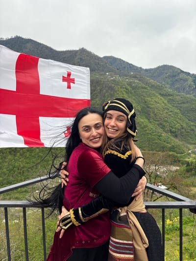 Two women embrace in front of a Georgian flag with a mountainous backdrop. One woman wears a black traditional Georgian headpiece and clothing with gold trim, while the other wears a red traditional garment with gold trim and has dark hair. They are standing on what appears to be a balcony with a metal railing. The background features green, tree-covered mountains under an overcast sky. The location is Kveda Chkhutuneti, Georgia.