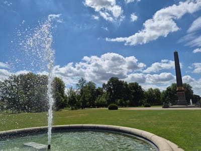 The image captures a sunlit scene in the Schloss (Palace) Park in Braunschweig, Germany, on a bright, cloudy day. In the foreground, a circular fountain with a stone rim is filled with rippling water, and a stream of water sprays upwards from its center. To the right of the fountain, a large, dark obelisk stands prominently, inscribed with text that is partially visible. The obelisk is surrounded by a low stone platform and other decorative elements, including what appear to be sculptures of lions at its base.
Beyond the fountain and obelisk, a vast expanse of manicured green lawn stretches into the background. A line of lush, mature trees forms a dense border along the middle ground, with more trees scattered throughout the park. The sky above is a vibrant blue, adorned with scattered white cumulus clouds, indicating fair weather. The overall impression is one of a peaceful, well-maintained urban park setting.