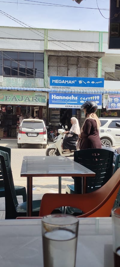 This street-level image depicts a vibrant commercial scene in Kotapantonlabu, Indonesia, during bright daylight hours under a clear sky. In the foreground, an outdoor seating area is visible, featuring a metal table, dark green, and orange plastic chairs, with an out-of-focus glass of water positioned prominently.
The midground captures daily street activity. A white Toyota Avanza with the license plate "BL 1171 KC" is parked, and two women are riding motorbikes. The woman closer to the foreground wears a dark brown top, a light hijab, and a black motorcycle helmet. Behind her, another woman in a light-colored hijab drives a white scooter.
The background reveals typical storefronts. A green sign advertises "TOKO KAIN FAJAR," a fabric shop that sells "PERLENGKAPAN BAYI, SEGALA JENIS KAIN SARUNG, BAD COVER, PERLENGKAPAN HAJI DAN UMRAH" (baby equipment, all types of fabric, sarongs, bed covers, Hajj and Umrah equipment). Adjacent to it are two blue signs for electrical retailers: "MEGAMAN® GERAI ELECTRIC," which mentions "Gen LED brand since 1994," and "Hannochs® GERAI ELEC," displaying various lightbulbs. Overhead, numerous power lines traverse the scene. The image captures the routine hustle of a commercial district, with elements of local culture and commerce.