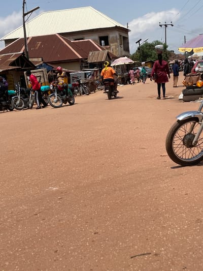 A ground-level, eye-level, wide shot captures a bustling street scene in Tangu, Nigeria, during daylight. The foreground is dominated by the dusty red earth of the street, interspersed with small white pebbles and occasional debris. In the mid-ground, several motorcycles are parked, some with riders, suggesting a local transport hub or market. People are actively engaged in various activities; one person in an orange shirt rides a motorcycle, while another, wearing a red top and black trousers, stands near a motorcycle. Further back, a woman walks with a large bowl balanced on her head, and other individuals are seen milling around market stalls and buildings. The background reveals a collection of structures with corrugated iron and white metal roofing, indicative of typical market architecture. The sky above is partly cloudy, casting a bright light on the scene. The presence of motorcycles and the general activity suggests this is a lively commercial area within Tangu.