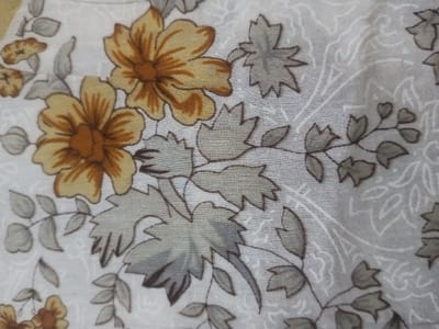 A floral textile features large yellow flowers with brown centers, accented by a pattern of grey and white leaves and vines. The background is a light, possibly off-white color with a faint, swirling pattern. The fabric has a textured appearance, likely linen or a similar material. The design creates a vintage or traditional aesthetic.