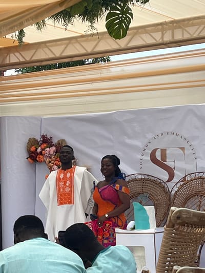 This image depicts a celebratory event in Kumasi, Ghana, likely a wedding or engagement reception. Two individuals, a man and a woman, stand prominently against a white banner as the focal point of the scene.
The man, positioned on the left, has dark skin and short dark hair. He wears a traditional white tunic with a central orange panel adorned with white intricate embroidery. He stands with his hands clasped behind his back, looking slightly to his left.
The woman, to his right, also has dark skin and dark hair styled upwards. She wears a vibrant, off-the-shoulder dress featuring an orange bodice, a pink and blue patterned overlay on the chest, and a multi-colored patterned skirt. A gold watch is visible on her left wrist, and she wears rings on her left hand. She is looking towards the camera with a slight smile.
Behind them, a white banner displays "SOLOMON AND EMMANUELLA" in a semi-circular arc above a large, stylized brown/gold "SE" logo. To the left of the banner, a floral arrangement incorporates orange, pink, and white flowers with dried palm-like elements. To the right, two large, round, wicker fan-like decorations are visible. A small white table stands to the right of the woman, holding a light blue, wave-shaped decorative object and a white flower.
In the immediate foreground, the backs of the heads and shoulders of three attendees are visible. They are wearing light blue collared shirts. The individual on the far right appears to be holding a dark-colored smartphone, directed towards the main subjects.
Above the scene, a light-colored tent or canopy structure with cream-colored fabric drapes and metal trusses is present. Artificial green foliage, including a large Monstera leaf, hangs from the overhead structure. Partially visible on the far right are elements of wicker chairs, indicating seating arrangements for guests. The lighting suggests an outdoor or semi-outdoor setting during daylight hours.