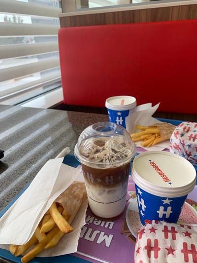 This media file features a close-up, angled view of a fast-food meal on a tray. The primary subjects are a drink in a clear plastic cup with a lid, a paper cup with the "HESBURGER" logo, and a serving of french fries in a paper wrapper. The drink appears to be iced coffee or a similar beverage with ice and milk. The french fries are golden brown and appear fresh.
The background shows a red booth or seating area, suggesting the setting is a restaurant or fast-food establishment. The tray itself has some purple branding with white text, part of which reads "MJI" and potentially other words that are obscured. The overall impression is a casual meal, likely taken during the day given the lighting. The image is oriented such that the food and drinks are the focus, with the red seating providing a contrasting color. No people are visible in the frame. The image does not provide explicit cues about the weather. The brand "HESBURGER" is clearly visible on one of the cups, indicating the likely origin of the food. The other cup and the wrapper for the fries also feature a repeating pattern of stylized "H" shapes and stars.