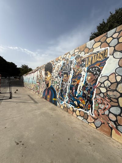 The image depicts a long, curved concrete wall adorned with vibrant graffiti art. The graffiti features stylized portraits of young men in soccer uniforms, with the words "LA MASIA" prominently displayed, indicating a connection to FC Barcelona's youth academy. The artwork is rendered in bold colors and dynamic poses, suggesting a celebration of soccer talent. The wall itself is textured with a pattern of colorful, irregularly shaped stones.

In the foreground, a concrete pathway runs alongside the wall, leading into a public space that appears to be a park or recreational area. To the left of the pathway, a soccer goal with black metal framing is visible. Two soccer balls rest on the ground near the goal. In the background, trees and a light blue sky with scattered clouds complete the scene. The overall impression is that of an outdoor urban environment, likely a public space where young people engage in sports and appreciate street art.

The location of this artwork is confirmed to be in the city of Mataró, Spain, specifically on the wall of the Parc de Can Mates. This park is known for its integration of public art and recreational facilities, making it a popular spot for both locals and visitors.