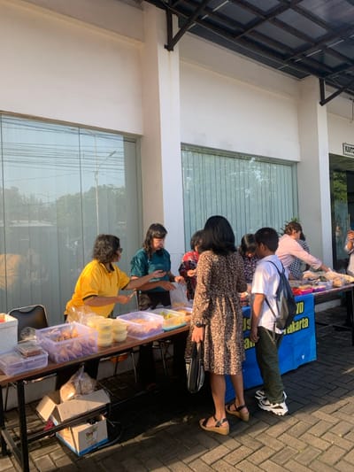 The scene takes place in front of a building located in Kecamatan Kartasura, Indonesia, at coordinates -7.567 latitude, 110.757 longitude. A group of people are gathered around a table displaying assorted food items in containers. A woman in a yellow shirt is interacting with a woman in a green shirt, while another woman wearing a brown floral dress stands nearby. A young boy wearing a backpack stands beside the table, observing the scene. The table is situated next to a large blue sign with the word "Jakarta" printed on it. The background features a building with large windows and a canopy overhead. The ground is a patterned brick surface, with a single black chair to the left of the table. The scene appears to be an outdoor market or food stall.