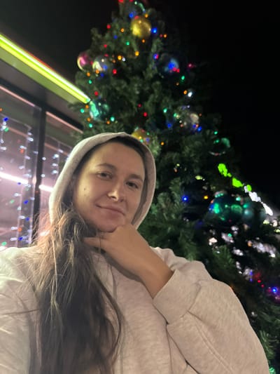 A woman poses in front of a Christmas tree. She wears a gray hoodie and rests her chin on her hand. Long brown hair falls across her shoulder. The Christmas tree is decorated with colorful lights and ornaments, with a window reflecting the lights. A yellow glow illuminates the left side of the image, contrasting the dark background. The picture is taken at a slight angle, creating a sense of casualness.