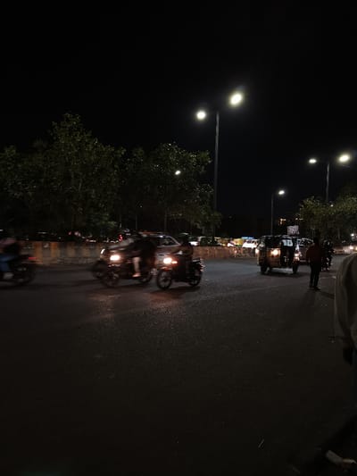 The image, taken at night in Noida, India (28.587, 77.375), shows a street with several vehicles passing by, lit by street lamps. There are three motorcycles traveling in the same direction, with the middle one closest to the camera. There are two people on the middle motorcycle. Behind the motorcycles, a small, white, three-wheeled vehicle is traveling on the same path. There are other cars, blurred and further away, as well as an individual on foot. The area appears to be relatively deserted.