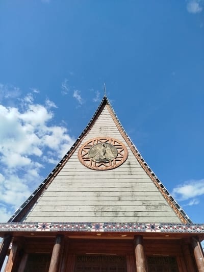 A low-angle shot shows the facade of a building in An Phu Ward, Vietnam. The building features a large, triangular roof with horizontal wooden planks, culminating in a thin, metallic point. A circular emblem adorns the center of the roof, decorated with a brick-like border. The emblem features a central figure, possibly human or angelic, though details are obscured. Decorative patterns of repeating geometric shapes run along the base of the roof. Supporting the roof are dark wooden pillars, suggesting an entrance or portico underneath. The sky forms the background, displaying a clear blue color with scattered, fluffy white clouds.