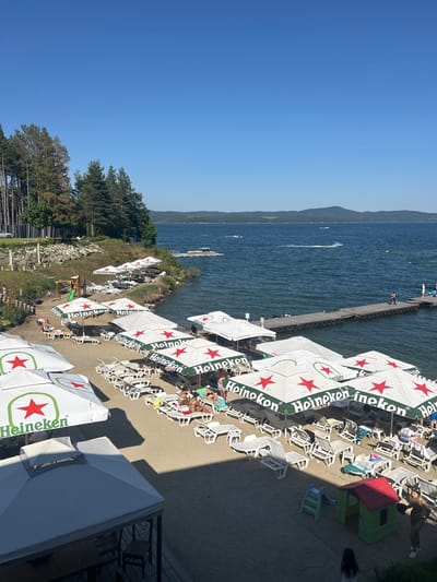 This is a high-angle, full shot of a beach resort on a sunny day, likely in Dolni Pasarel, Bulgaria, overlooking a large body of water. The foreground shows a sandy beach populated with numerous white lounge chairs and several large umbrellas, many branded with "Heineken" logos. Some people are visible on the beach, either lying on lounge chairs or walking.
Further back, a wooden pier extends into the water, where a few individuals are standing or walking. Several boats, including jet skis and a larger watercraft, are present on the lake, creating wakes. The water is a deep blue and appears calm, with a distant treeline and rolling hills forming the horizon under a clear blue sky. To the left of the beach, a grassy slope is covered with pine trees, and a small playground with a slide is visible. Structures resembling covered seating areas are also present in the foreground. The scene depicts a typical summer recreational setting at a lakeside resort.