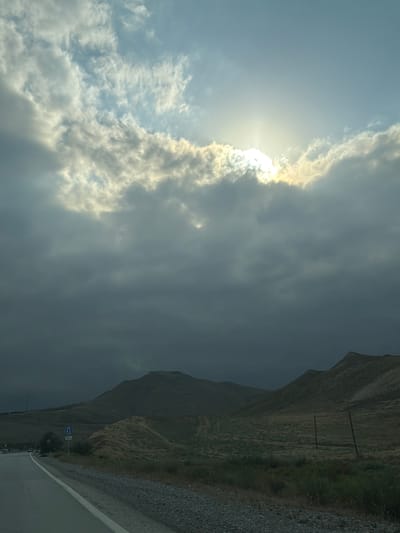 The image depicts a landscape with a road in the foreground, leading into rolling hills and mountains under a dramatic sky. The sun is partially obscured by thick clouds, casting rays of light through the overcast. A traffic sign with a blue "U-turn" symbol and "300m" is visible on the right side of the road. The terrain appears arid and sparsely vegetated, with dry grass and some scattered bushes. The overall impression is one of a vast, open natural environment with a road suggesting human passage. The location is identified as Jangi, Azerbaijan.