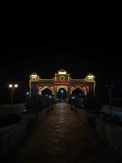 A night view of the entrance archway of a park or memorial in Suzak, Kyrgyzstan, is depicted. The archway is illuminated with bright yellow and red neon lights, highlighting its intricate architectural details and calligraphy in Cyrillic script. The pathway leading to the arch is paved with textured tiles, reflecting the light from the illumination and streetlights. Several trees are planted along the sides of the pathway and in front of the archway, with some benches visible. Two traditional-style street lamps are positioned on either side of the pathway in the foreground. The overall scene is dark with the exception of the illuminated structures and pathway, suggesting a nighttime setting.