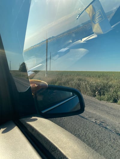 A hand with tan skin is seen through a car window, touching the window. The hand is positioned in front of a rural landscape outside the car, with a field of sunflowers and a dirt road visible. A side-view mirror of the car is also in the frame. A Ukrainian flag is attached to the car's dashboard, visible through the windshield. The scene is set in Maiske, Ukraine, under a clear blue sky.