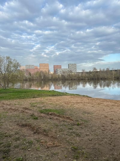 The image presents a view of Gavrilkovo, Russia. The scene is dominated by reflections in a body of water. Reflected are several high-rise apartment buildings, some clad in light-colored panels and others in a reddish-brown material. A line of trees is also reflected in the water. The sky above is filled with layered, textured clouds, also reflected in the still water. The foreground features a bank composed of dirt and sparse grass. In the lower left, a tree is partially visible. The photograph appears to be taken at an angle or perhaps is inverted.