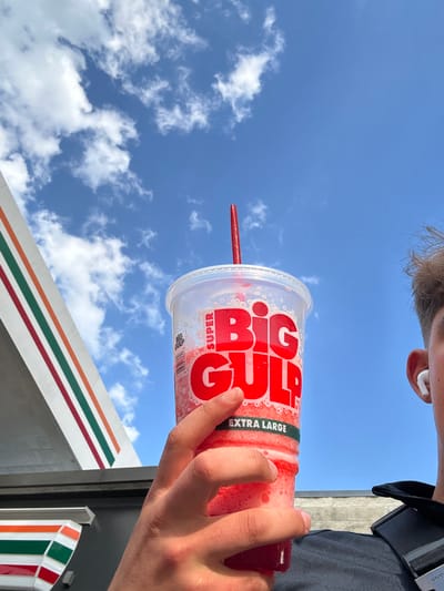 A person holds a large "Big Gulp" cup filled with a red beverage in front of a 7-Eleven store on a sunny day. The cup features the red "BIG GULP" logo with the words "SUPER" and "EXTRA LARGE." A red straw is visible. The person is wearing an AirPod and a dark-colored shirt. A glimpse of the 7-Eleven's roofline and logo are visible. The location of the 7-Eleven is presumably Tampa, United States. The sky is blue with scattered clouds.